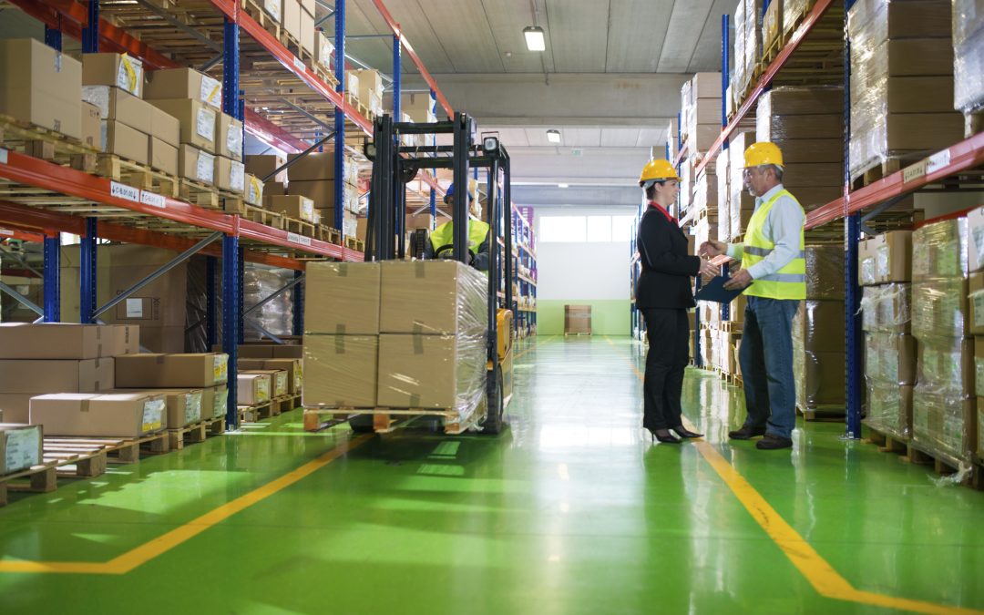 A warehouse with a forklift on the left and a man and woman speaking with hard hats on.